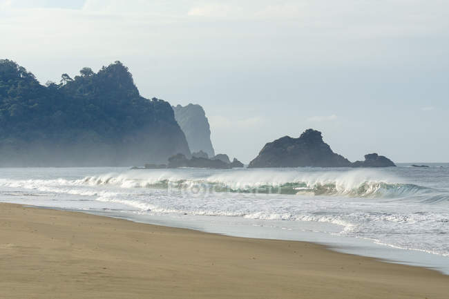 Indonesia, Java Timur, Kabany Banyuwangi, Parque Nacional Meru Betiri, olas en la playa solitaria, siluetas de rocas escénicas en el fondo - foto de stock