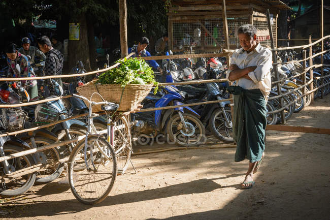 Велосипед с корзиной овощей и человек, проходящий возле мопедов парковка на фермерском рынке, Nyaung-U, Мандалайская область, Мьянма — стоковое фото
