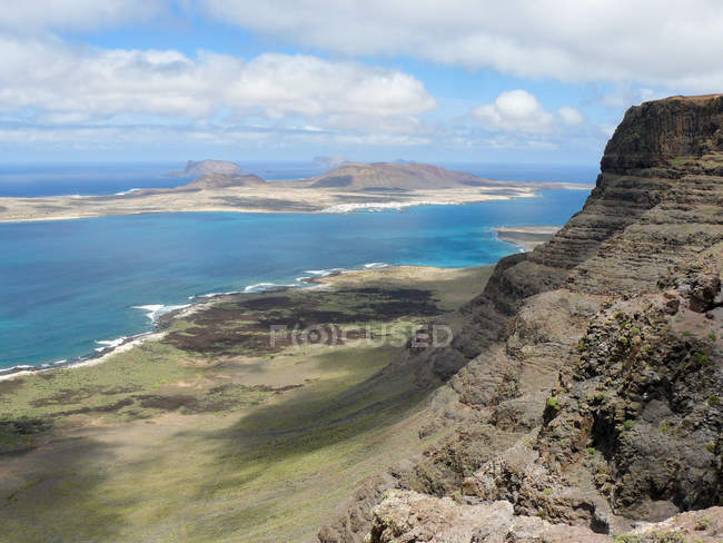 España, Islas Canarias, Teguise, acantilados del macizo de Famara e isla costera de La Graciosa con la localidad de Caleta del Sebo desde arriba - foto de stock