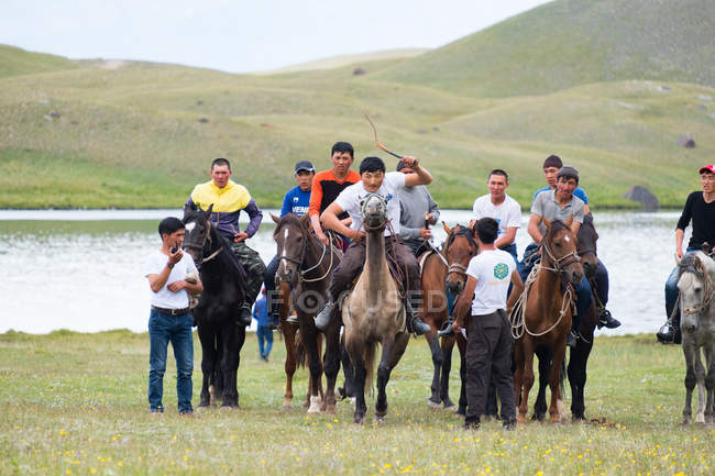 Región OSH, KYRGYZSTAN - 22 de julio de 2017: Juegos nómadas, hombres a caballo, paisaje de montaña con lago en el fondo - foto de stock