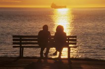 Siluetas de pareja en el banquillo disfrutando de la puesta de sol en Stanley Park, English Bay, Vancouver, British Columbia, Canadá - foto de stock