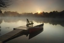 Salida del sol sobre canoa atada al muelle con silla en Muskoka, Ontario, Canadá - foto de stock