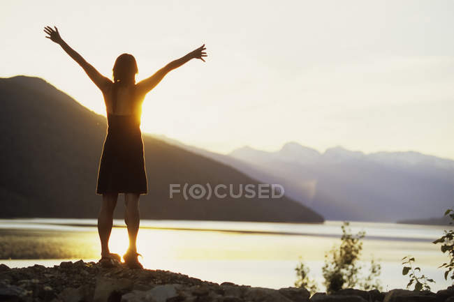 Vista trasera de la mujer con los brazos extendidos en la orilla del río Columbia cerca de Revelstoke, Columbia Británica, Canadá . - foto de stock