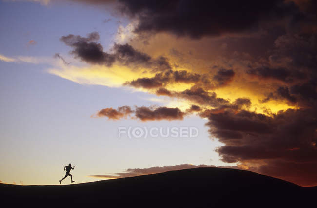 Силуэт женщины, бегущей по хребту в сторону шторма на закате, Национальный парк Каньонлендс, Моаб, Юта, США — стоковое фото
