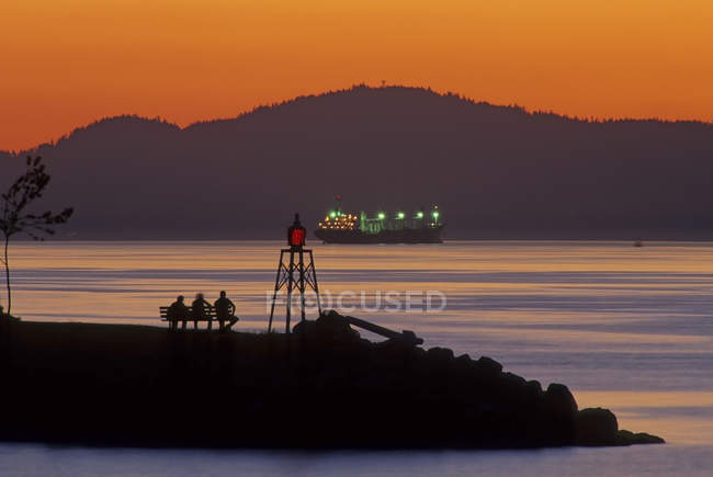 Silhuetas de pessoas em exibição de banco por do sol com cargueiro iluminado, Burrard Inlet, Vancouver, British Columbia, Canadá — Fotografia de Stock