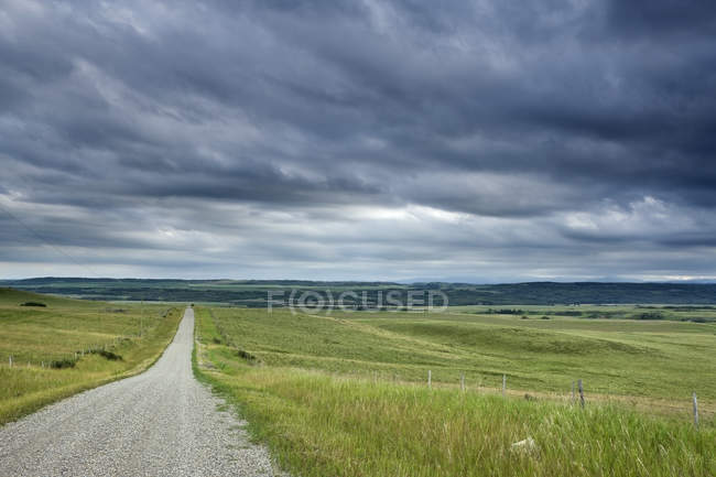 Strada di campagna attraverso il campo e la recinzione sotto le nuvole di tempesta vicino a Cochrane, Alberta, Canada — Foto stock