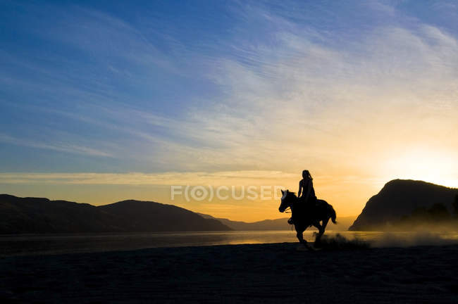 Женщина наслаждается бегом с лошадью после заката на берегу реки Томпсон, Камлупс, Британская Колумбия, Канада — стоковое фото