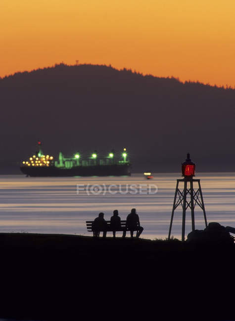 Siluetas de personas en el banco viendo atardecer con carguero iluminado, Burrard Inlet, Vancouver, Columbia Británica, Canadá - foto de stock