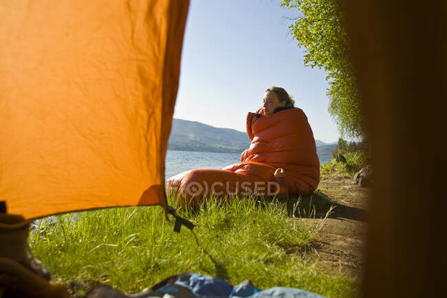 Giovane donna seduta avvolta nel sacco a pelo vicino alla tenda, Skaha Lake, Penticton, British Columbia, Canada — Foto stock