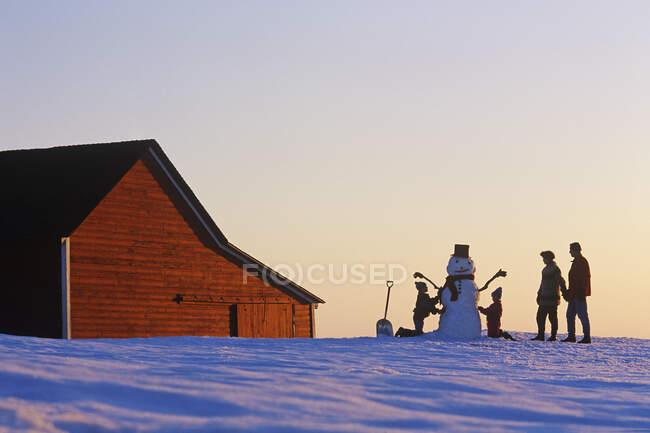 Una familia hace un muñeco de nieve frente a un granero rojo, cerca de Glass, Manitoba, Canadá - foto de stock
