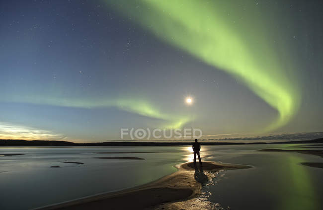 Person standing along shore of MacKenzie River with aurora borealis overhead, Northwest Territories, Canada. — Stock Photo
