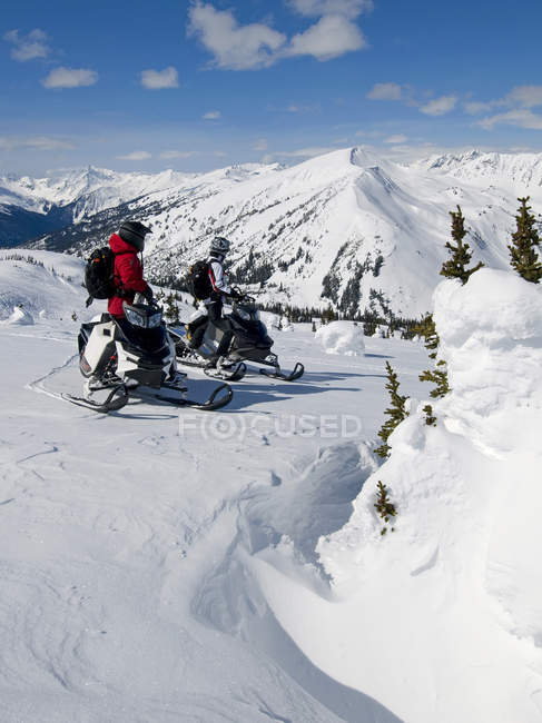 Amigos se detienen y miran a la vista mientras motos de nieve, montañas Monashee, Valemount, Thompson Okanagan, Columbia Británica, Canadá - foto de stock