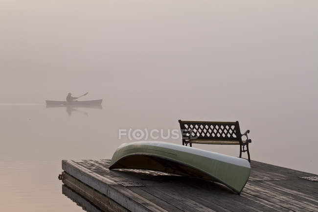 Doca com banco e barco e silhueta de homem remando canoa em Oxtongue Lake, Muskoka, Ontário . — Fotografia de Stock