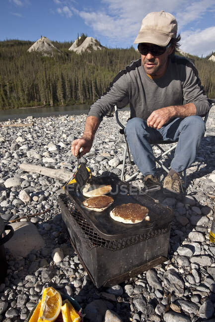 MIddle-aged man cooking pancakes over firebox while camped on Nahanni River, Nahanni National Park Preserve, NWT, Canada. — Stock Photo