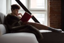 Schöne Frau liest Buch, während sie es sich auf dem Sofa im Wohnzimmer gemütlich macht — Stockfoto
