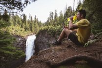 Uomo seduto vicino alla cascata e acqua potabile in una giornata di sole — Foto stock