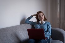 Задумчивая женщина с ноутбуком сидит на диване в гостиной на дому — стоковое фото