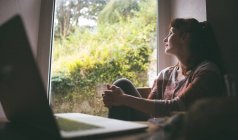 Задумчивая женщина сидит у окна дома — стоковое фото
