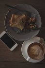 Sobrecarga de taza de capuchino con envoltura de alimentos y teléfono móvil en la mesa - foto de stock