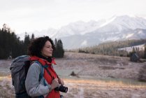 Задумчивая женщина, стоящая с рюкзаком и цифровой камерой зимой — стоковое фото
