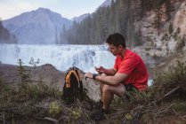 Молодой человек использует мобильный телефон возле водопада — стоковое фото