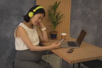 Беременная деловая женщина слушает музыку на мобильном телефоне за столом в офисе — стоковое фото