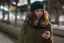 Thoughtful woman in winter clothing holding mobile phone in platform — Stock Photo