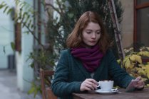 Mujer usando teléfono móvil mientras tiene cappuccino en la cafetería al aire libre - foto de stock