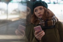 Thoughtful woman in winter clothing leaning on glass wall at railway station — Stock Photo