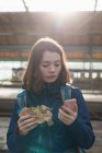 Mujer usando teléfono móvil mientras tiene comida envuelta en la estación de tren - foto de stock