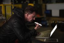 Механик разговаривает по мобильному телефону во время использования ноутбука в гараже — стоковое фото