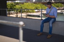 Чоловік використовує мобільний телефон, сидячи на паркані в сонячний день — стокове фото