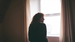 Вид ззаду вдумливої жінки, що стоїть біля вікна вдома — стокове фото