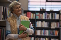Mujer mayor reflexiva sosteniendo un libro en la biblioteca - foto de stock