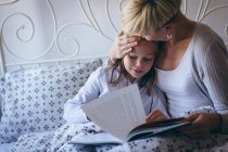 Любящая мать целует свою дочь во время чтения книги в спальне — стоковое фото