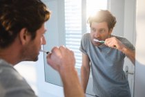 Мужчина смотрит в зеркало и чистит зубы в ванной дома — стоковое фото