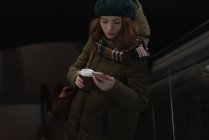 Mulher com xícara de café usando telefone celular na escada rolante na estação ferroviária — Fotografia de Stock