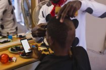 Cliente que usa el teléfono móvil mientras el peluquero se corta el pelo en la peluquería - foto de stock