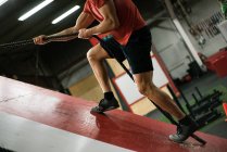 Homem musculoso escalando uma parede inclinada com corda no ginásio — Fotografia de Stock