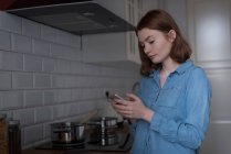 Молода жінка використовує мобільний телефон на кухні — стокове фото