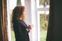 Mulher atenciosa olhando pela janela enquanto toma uma xícara de café em casa — Fotografia de Stock