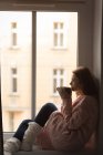 Frau trinkt Kaffee, während sie zu Hause durchs Fenster schaut — Stockfoto