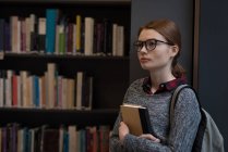 Thoughtful woman with book standing in the library — Stock Photo