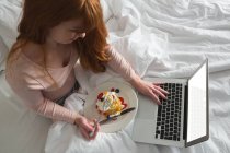 Mujer de pelo rojo usando portátil en el dormitorio con postre en el plato - foto de stock