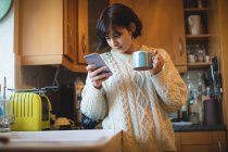 Mujer tomando café mientras usa el teléfono móvil en la cocina en casa - foto de stock