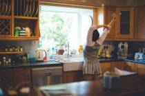 Visão traseira da mulher de pé com os braços para cima na cozinha em casa — Fotografia de Stock