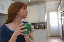 Mujer pensativa tomando café en taza verde en casa - foto de stock