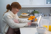 Capelli rossi donna sorridente utilizzando il computer portatile in cucina a casa — Foto stock