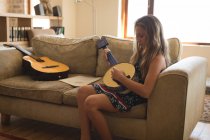 Девушка играет на банджо-гитаре в гостиной дома и сидит на диване — стоковое фото
