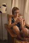 Дівчина використовує мобільний, сидячи на стільці у кімнаті біля вікна та лампи — стокове фото